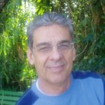 homem branco, de meia idade, cabelo grisalho, curto e farto, sorri sutilmente, olhando para frente. Ele usa óculos e uma camiseta azul claro e escuro. Ao fundo, uma vegetação verde.