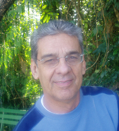 homem branco, de meia idade, cabelo grisalho, curto e farto, sorri sutilmente, olhando para frente. Ele usa óculos e uma camiseta azul claro e escuro. Ao fundo, uma vegetação verde.
