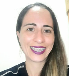 Selfie de uma mulher de aparelho em seus 40 anos sorrindo. Seu cabelo castanho com pontas loiras está colocado sobre seu ombro esquerdo, descendo até o busto. Ela usa uma blusa preta e batom roxo.