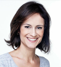 Mulher de 30 anos retratada do busto para cima sorrindo. Seu cabelo, no estilo chanel, é preto com finas mechas claras. Ela usa uma blusa cinza. Atrás dela, há um fundo branco.