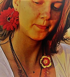 Retrato de uma jovem mulher do busto até a testa. O sol ilumina o lado direito do seu rosto. A jovem de cabelos loiros chanel usa um brinco grande em forma de flor vermelha. Sobre seu pescoço, dois colares: um preto e outro com flores.