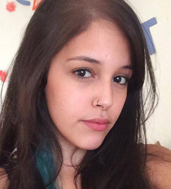 Selfie de uma jovem mulher de cabelos longos com uma mecha azul. O cabelo liso escorre pelos dois ombros. Ela tem um piercing preto na parte direita do nariz. Seus olhos são pretos e refletem um pequeno círculo de luz fundo.