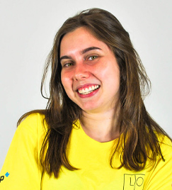 Uma jovem de 20 anos retratada em fundo branco. Seus cabelos castanhos claros ultrapassando seu ombro, cobrindo parte de sua camiseta amarela. Ela sorri, inclinando o rosto para a direita. Sua bochecha está avermelhada.