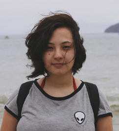Uma jovem mulher retratada do busto para cima em uma praia. Ela veste uma camiseta cinza com a figura de um alienígena. O vento sopra seu cabelo liso e preto na altura do queixo. Embaixo de seu olho direito, há uma pinta circular. Ao fundo, o mar de horizonte.