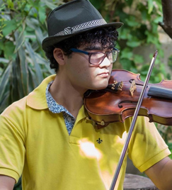 Um jovem de óculos toca violino. Com a mão esquerda, ele segura o instrumento e, com a esquerda, o arco. Ele veste uma camiseta polo amarela, calça jeans e um chapéu preto. Atrás dele, muitas árvores.