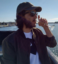 Um jovem de 20 anos bebendo uma taça de champanhe. Ele está de pé em um barco, vestindo boné, óculos escuros e uma jaqueta bege. Ao fundo, um rio se estende e reflete o sol em vários pontos.