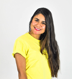 Retrato de uma jovem mulher da cintura para cima. Vestindo uma camiseta amarela com a manga dobrada, ela inclina seu ombro direito para frente. Todo seu cabelo liso e castanho com mechas loiras está colocado sobre o ombro esquerdo. Suas bochechas arredondadas refletem uma luz externa.
