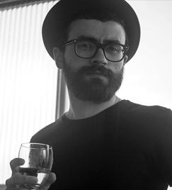 Selfie em preto e branco de um jovem segurando uma taça com a mão direita. Ele veste um chapéu redondo, óculos pretos e uma camiseta preta simples. Seu rosto é recoberto por barba volumosa. Ao fundo, persianas ilunimadas.