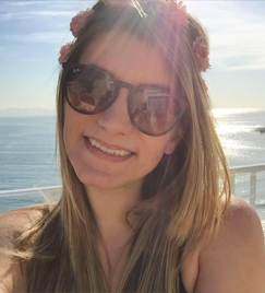 Selfie de uma jovem mulher loira iluminada pelo sol. Ela usa óculos escuros e sorri para a foto. No topo de sua cabeça, há uma tiara de flores entrelaçada com seu cabelo liso e longo. Ao fundo, aparece o mar refletindo raios do sol.