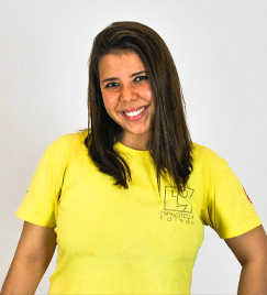 Jovem mulher, com cabelos pretos lisos na altura do ombro, posa para foto. Vestindo uma camiseta amarela de mangas curtas, ela aparece da cintura para cima, com os braços abaixados.