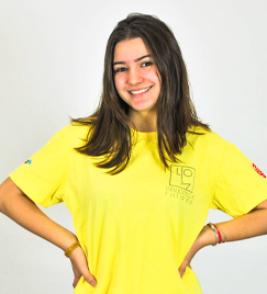 Uma jovem mulher leva suas mãos ao quadril e sorri. Em um fundo branco, ela aparece do tronco para cima. Seus cabelos lisos e escuros escorrem pelo seu pescoço até os ombros, fazendo contraste com a camiseta amarela que veste.