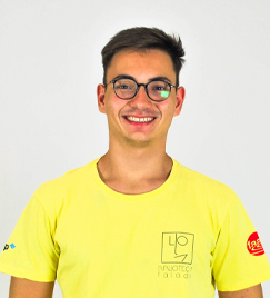 Um jovem em seus vinte anos, de camiseta amarela, posa para uma foto. Seu cabelo é mais volumoso em cima e curto nos lados. Ele usa óculos pretos, que refletem uma pequena luz.