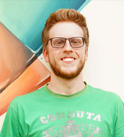 Do peito para cima, um jovem vestindo uma camiseta verde olha para frente e sorri. Ele tem barba, cabelo, sobrancelhas e cílios loiros. Em seu rosto, há também um óculos retangular. Ao fundo, aparece uma pintura com formas geométricas em verde e laranja.