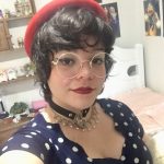 Uma moça branca de cabelos pretos e escuros, usa óculos e está usando um batom vermelho escuro. Está vestindo um vestido escuro com bolinhas brancas e em seu pescoço ela usa vários colares juntamente com uma gargantilha de coração. Ela está dando um leve sorriso para a foto.
