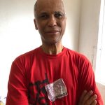 Foto de Ednilson Sacramento, homem negro de 50 anos, sem cabelos, rosto fino, sobrancelhas finas, braços cruzados, camiseta vermelha. Parede branca ao fundo.