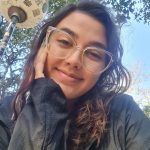 Fotografia de Laura estilo selfie. Laura, uma jovem mulher branca com cabelos castanhos claros, olha e sorri para a câmera. Ela usa óculos, tem sobrancelhas grossas e olhos castanhos, com um piercing na narina esquerda. Laura traja uma camiseta branca e uma jaqueta preta. Ao fundo, o céu azul e o topo de árvores.