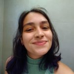Fotografia frontal de Barbara, jovem branca de 20 anos, cabelo castanho escuro com mechas roxas nas pontas. Sorri para a câmera com sobrancelhas grossas e três brincos na orelha esquerda. Veste camisa de gola alta sem mangas verde claro, fundo com parede cinza e piso.