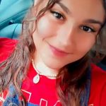 Fotografia estilo selfie de Bianca, jovem branca de 20 anos com cabelos ondulados castanhos claros. Ela sorri com delineador preto, batom rosa claro e colar dourado. Veste camiseta vermelha e macacão jeans, com o interior de um carro ao fundo.