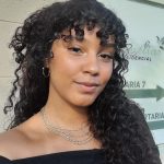 Uma mulher jovem sorri sem mostrar os dentes. Ela tem cabelos cacheados pretos na altura do peito e olha para a câmera. Está com brincos de argola e uma corrente com três tiras douradas no pescoço. Usa um vestido preto de decote estilo ombro a ombro. Atrás dela, há uma parede verde.