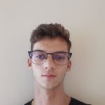 A fotografia de um homem jovem de cabelos curtos castanhos claros com expressão séria para a câmera. Ele veste uma camiseta preta e óculos de armação da mesma cor. Atrás dele, há uma parede branca.