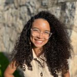 Fotografia de busto para cima de uma jovem mulher parda. Ela tem cabelos cacheados castanhos, sorri e está iluminada pelo sol. Usa óculos arredondados com armação transparente e um par de brincos com miçangas. Ela veste um macacão bege sem mangas. Ao fundo, há uma parede de pedras acinzentadas.