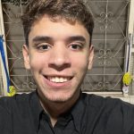 Selfie de um jovem de cabelo e olhos castanhos. Ele sorri para a foto e usa camisa social preta. Atrás dele, há uma janela aberta com uma grade branca.