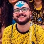 No centro da imagem, um jovem branco careca vestido com uma camiseta amarela com os escritos NAUMTERIA em preto. Ele tem barba e bigode, usa um óculos retangular com uma armação preta e está sorrindo. No topo de sua cabeça, há o desenho de uma seta azul apontada para baixo.