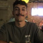 Foto de um rapaz de bigode e cabelo curto cacheado sorri sem mostrar os dentes. Ele usa uma camiseta cinza e, atrás dele, há uma parede revestida de tijolos aparentes.