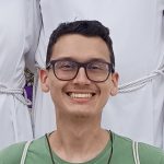 Um rapaz branco, de cabelo castanho escuro e um singelo topete sorri para a foto. Ele usa óculos e uma camiseta verde. Atrás dele aparecem tecidos brancos, das roupas de outras pessoas.