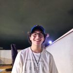 Fotografia de um homem jovem sorrindo com cabelo longo na cor preta. Ele veste uma camisa branca, um boné azul e usa um óculos com armação preta. No fundo, há uma parede branca com uma lâmpada de luz forte. O céu está nublado e escuro.