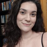 Selfie de uma mulher branca com rosto oval, de cabelos castanhos, compridos e ondulados. Ela sorri levemente para a câmera. Ela usa uma regata preta e possui um piercing prateado no septo. Atrás dela há uma estante cheia de livros.