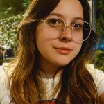 Selfie de uma jovem branca, com cabelos castanhos na altura um pouco abaixo dos ombros. Ela sorri discretamente para a foto. A jovem usa óculos redondos de armação fina. Ela veste uma camiseta branca com a estampa da mulher maravilha. Ao fundo da foto, há luzes penduradas em árvores durante a noite.