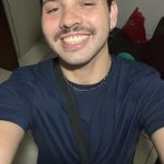 Fotografia em estilo selfie de um jovem branco. Ele tem cabelo encaracolado e castanho escuros, bigode e um piercing na sobrancelha direita. Ele usa um colar cinza, uma camiseta azul escura e uma shoulder bag.