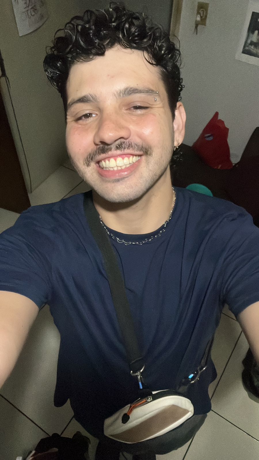 Fotografia em estilo selfie de um jovem branco. Ele tem cabelo encaracolado e castanho escuros, bigode e um piercing na sobrancelha direita. Ele usa um colar cinza, uma camiseta azul escura e uma shoulder bag.