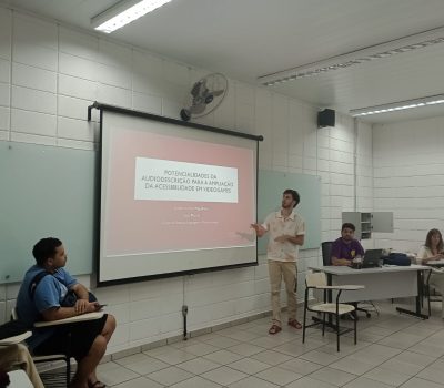 A foto mostra ao centro o palestrante em pé olhando em direção aos slides projetados na parede, ao redor há alunos sentados olhando para o palestrante.
