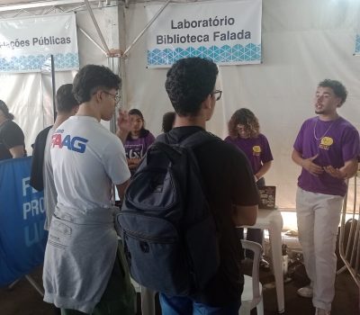 Fotografia de estudantes no estande do Laboratório Biblioteca Falada na Feira de Profissões UNESP 2024. Membros do laboratório estão em pé atrás de mesas de plástico brancas. De costas para a câmera e de frente para as mesas, estão três alunos. Ao fundo, há uma lona branca e um banner escrito "Laboratório Biblioteca Falada".