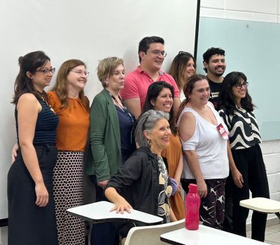 Fotografia lateral de um grupo de pessoas reunidas. Eles estão em uma sala de aula. Eles sorriem para a foto. Ao fundo há uma lousa branca.