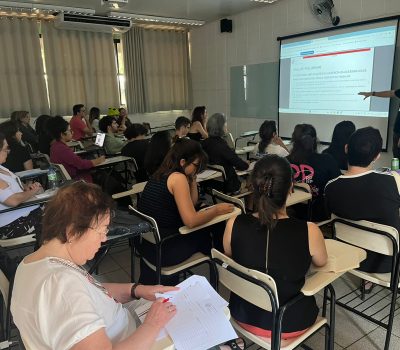 Fotografia de um grupo de pessoas sentadas em cadeiras. Elas estão em uma sala de aula. Ao fundo há um homem que aponta para uma apresentação de slides projetado na parede.