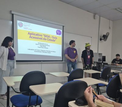 Fotografia de três pessoas em uma sala de aula. Eles estão ao lado de uma tela de apresentação de slides. Do lado direito do slide estão dois homens. Do lado esquerdo do slide está uma mulher. Todos estão uniformizados com a camiseta roxa do Biblioteca Falada. No slide está escrito: "Aplicativo SIGA: Guia Acessível da Cidade". Em frente aos membros do Biblioteca falada estão algumas carteiras escolares vazias.