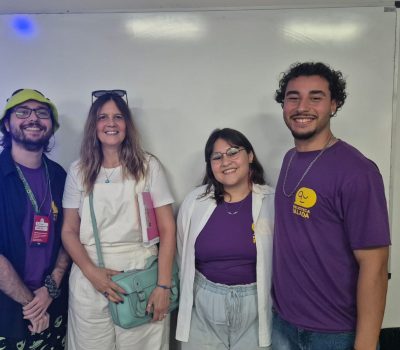 Fotografia de um grupo de pessoas sentadas em cadeiras. Elas estão em uma sala de aula. Ao fundo há um homem que aponta para uma apresentação de slides projetado na parede.