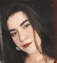 Foto de uma mulher branca que olha para a frente com a cabeça levemente inclinada para a esquerda. O cabelo dela é castanho escuro, liso e na altura dos ombros. Ela possui lábios finos com um pequeno sorriso, e está usando um batom vermelho escuro. Na orelha esquerda, é possível ver parte de um brinco prata.
