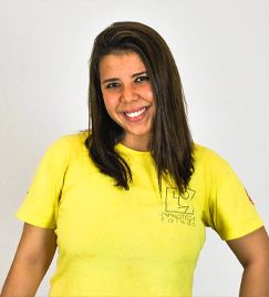 Jovem mulher, com cabelos pretos lisos na altura do ombro, posa para foto. Vestindo uma camiseta amarela de mangas curtas, ela aparece da cintura para cima, com os braços abaixados.