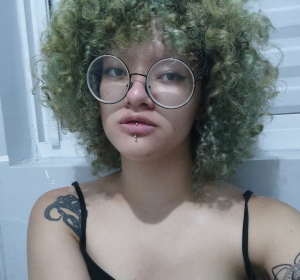 Melissa, jovem de pele clara, cabelos cacheados verde claro, óculos redondos. Piercing no lábio. Tatuagens de polvo e aranha. Blusa preta, plano frontal. Parede branca de fundo.