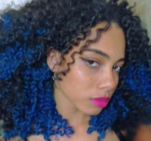 Fotografia de perfil estilo selfie de Heloísa, jovem negra de cerca de 20 anos. Cabelo cacheado tingido de azul, sobrancelhas finas, batom rosa nos lábios. Brinco de pedraria azul, blusa de alça preta. Fundo com parede branca.