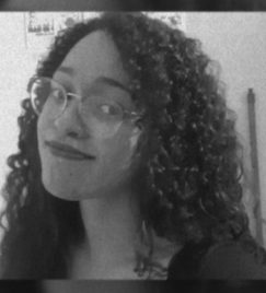Fotografia em filtro cinza do rosto de Ana Laura, jovem negra de 20 anos, cabelo cacheado escuro, sorri para a câmera com óculos de armação prateada. Usa batom vermelho e blusa preta, com fundo de parede branca.