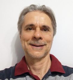 homem branco, de meia idade, rosto fino, cabelos brancos lisos e sobrancelhas pretas, sorri. Ele veste uma camiseta polo nas cores vinho, azul e cinza. Uma barba bem rasa cobre a parte inferior do rosto.