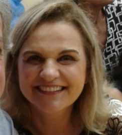 mulher branca, de meia idade, rosto arredondado, sorri. Com cabelos loiros e lisos emoldurando seu rosto até os ombros, sua figura ocupa a maior parte da tela