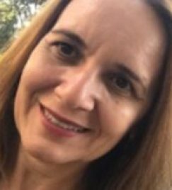 Mulher branca de meia idade, cabelos castanhos claros, lisos e longos. Ela tem olhos castanhos escuros e está sorrindo. Usa batom rosado.