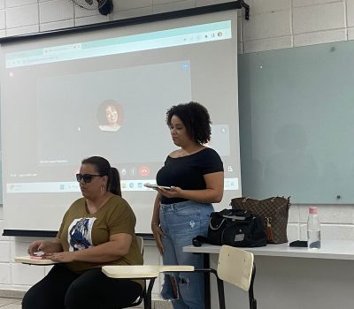Fotografia de Meirellen Godoi, mulher com deficiência visual, apresentando o seminário. Ao seu lado, está uma estudante, Meireleen está sentada enquanto apresenta. Ao fundo, está uma parede branca e uma tela onde uma ligação do google meet está sendo projetada.