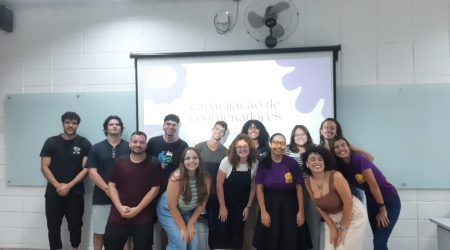 Foto do grupo de coordenadores do Biblioteca Falada, são treze pessoas em uma sala de aula. A maioria está de pé e alguns agachados, estão todos direcionados para frente e grande parte está sorrindo. Há uma projeção de slides ao fundo sobrepondo uma lousa de vidro. Ao fundo há uma parede branca.